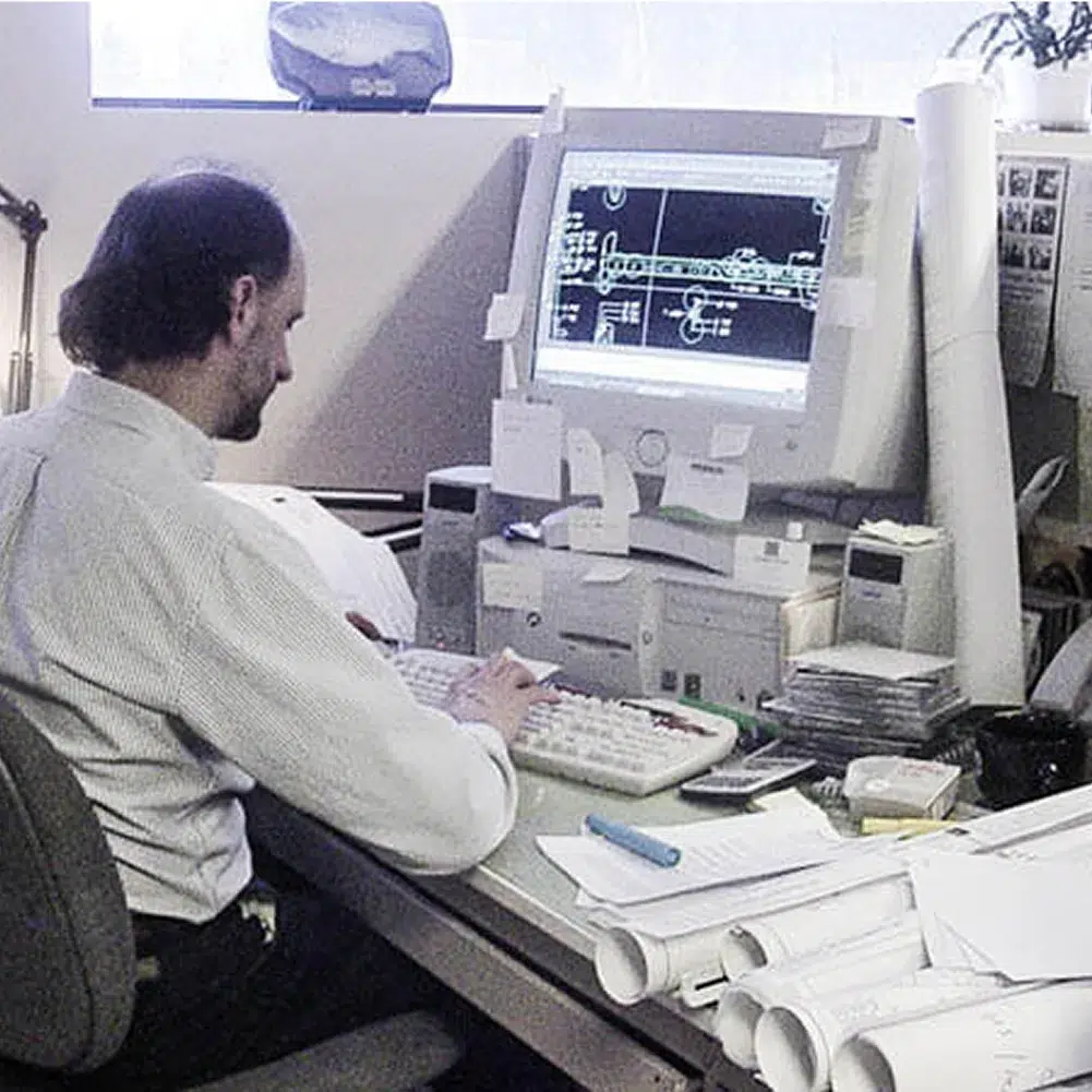 A man sits at a cluttered desk in a Maine engineering firm, working on technical drawings amid papers, office supplies, and rolled-up blueprints.