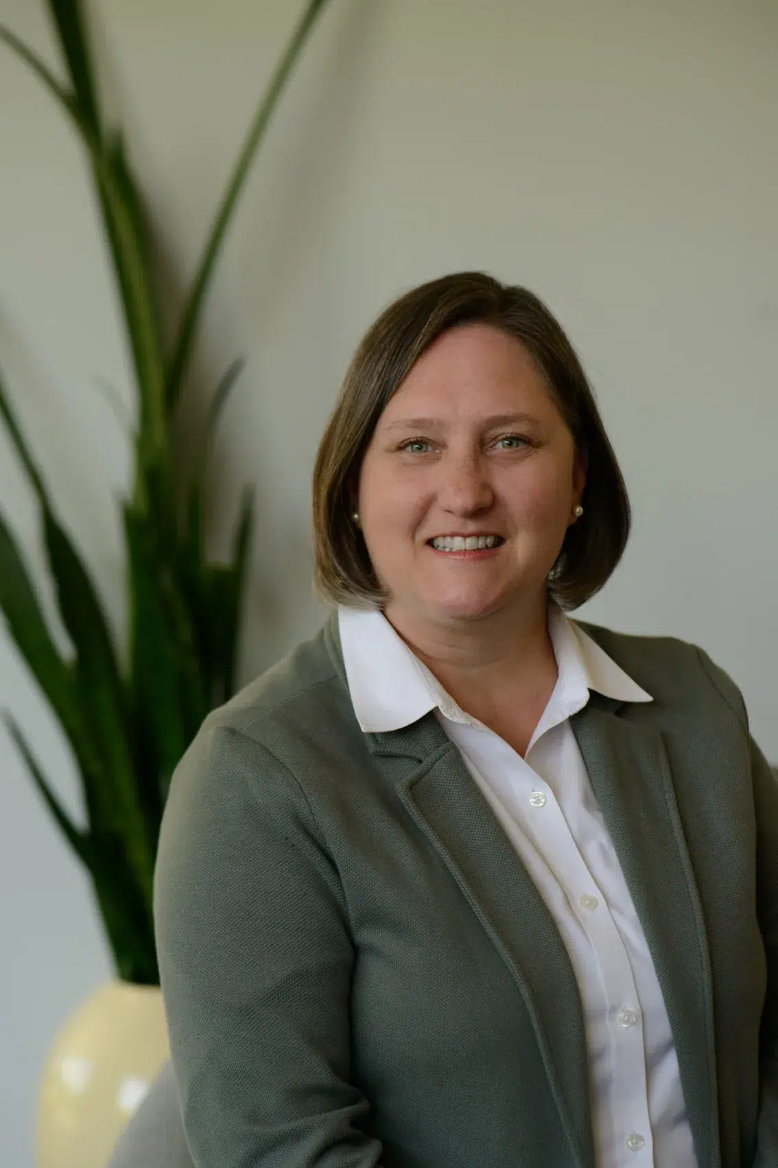 A woman with short brown hair wearing a white shirt and gray blazer smiles while sitting indoors near a tall green plant.