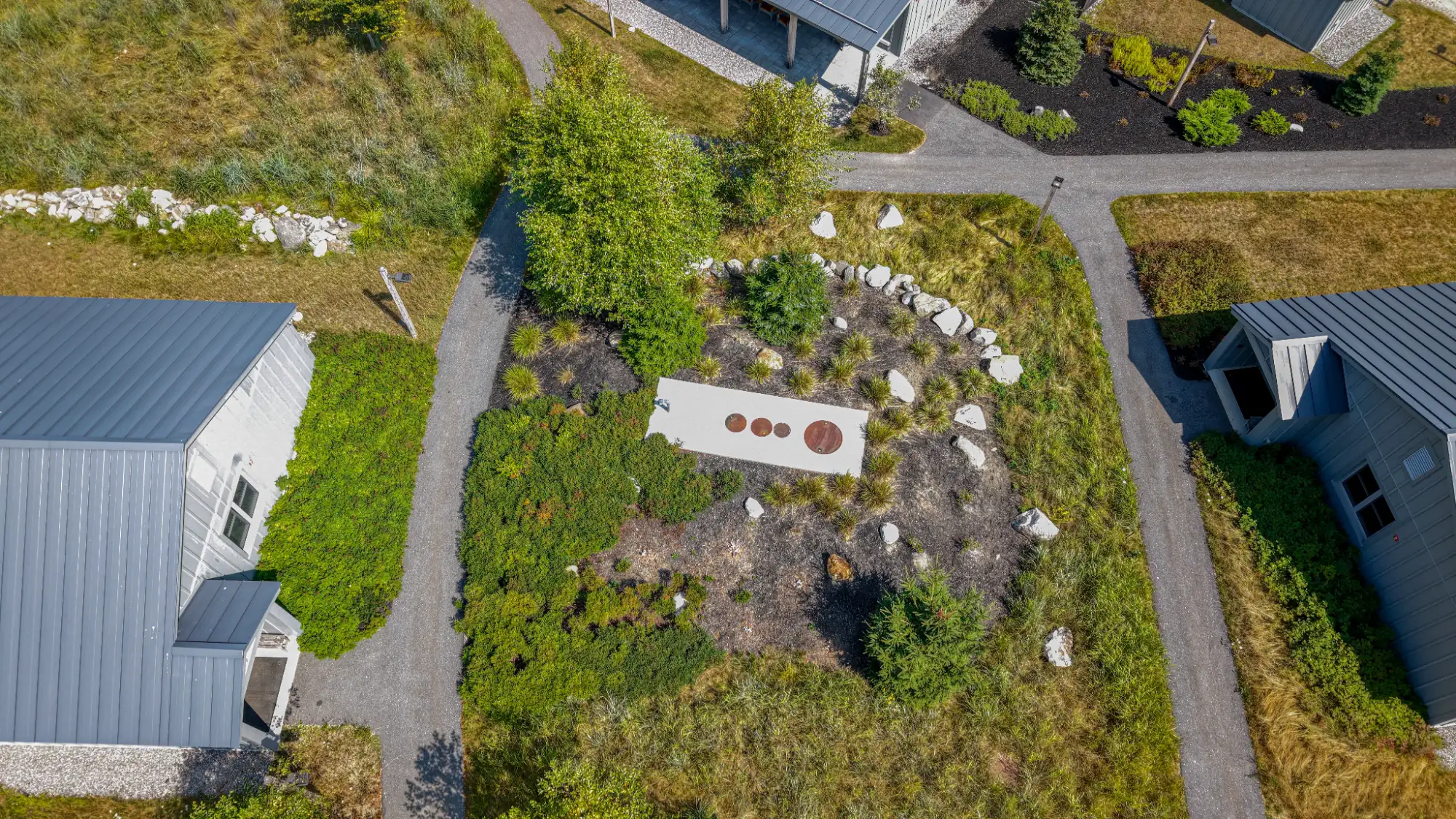 Aerial view of a landscaped garden designed with civil engineering services, featuring a white rectangular path, circular sculptures, rocks, and lush plants between two modern buildings.