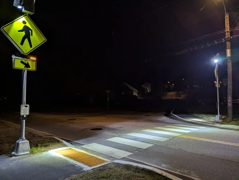 A crosswalk at night is lit by streetlights, with illuminated pedestrian crossing signs and a tactile paving strip—showcasing thoughtful traffic engineering.
