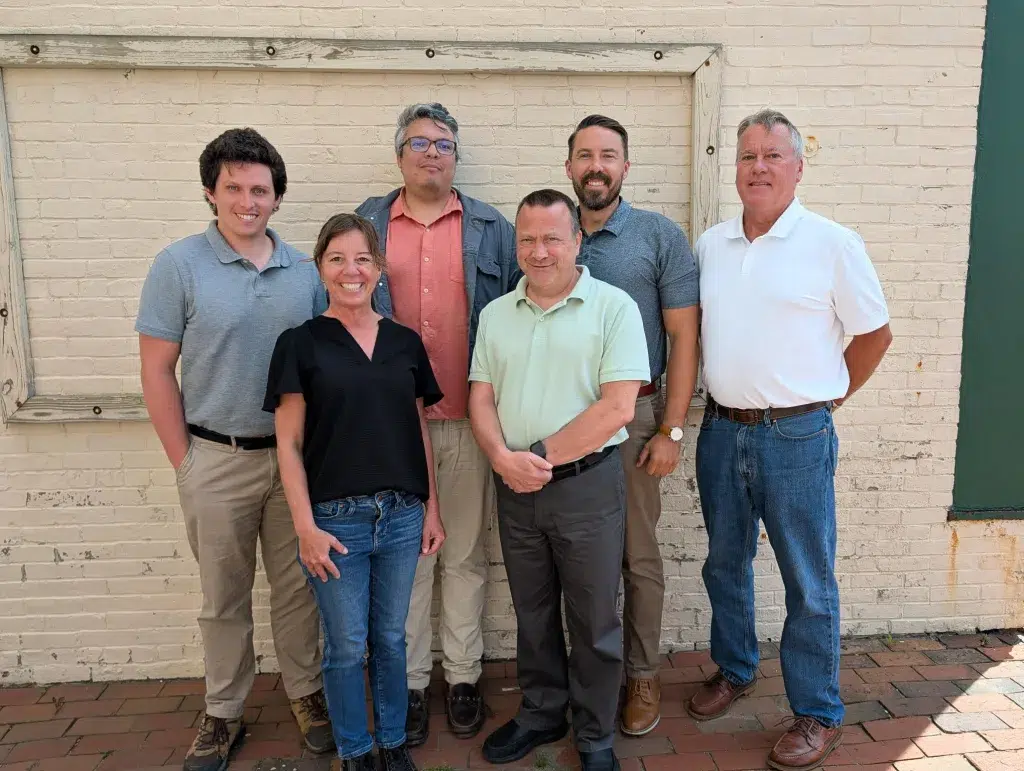 Seven adults posing together outdoors in front of a light brick wall with a wooden frame. They wear casual shirts and jeans and smile at the camera.