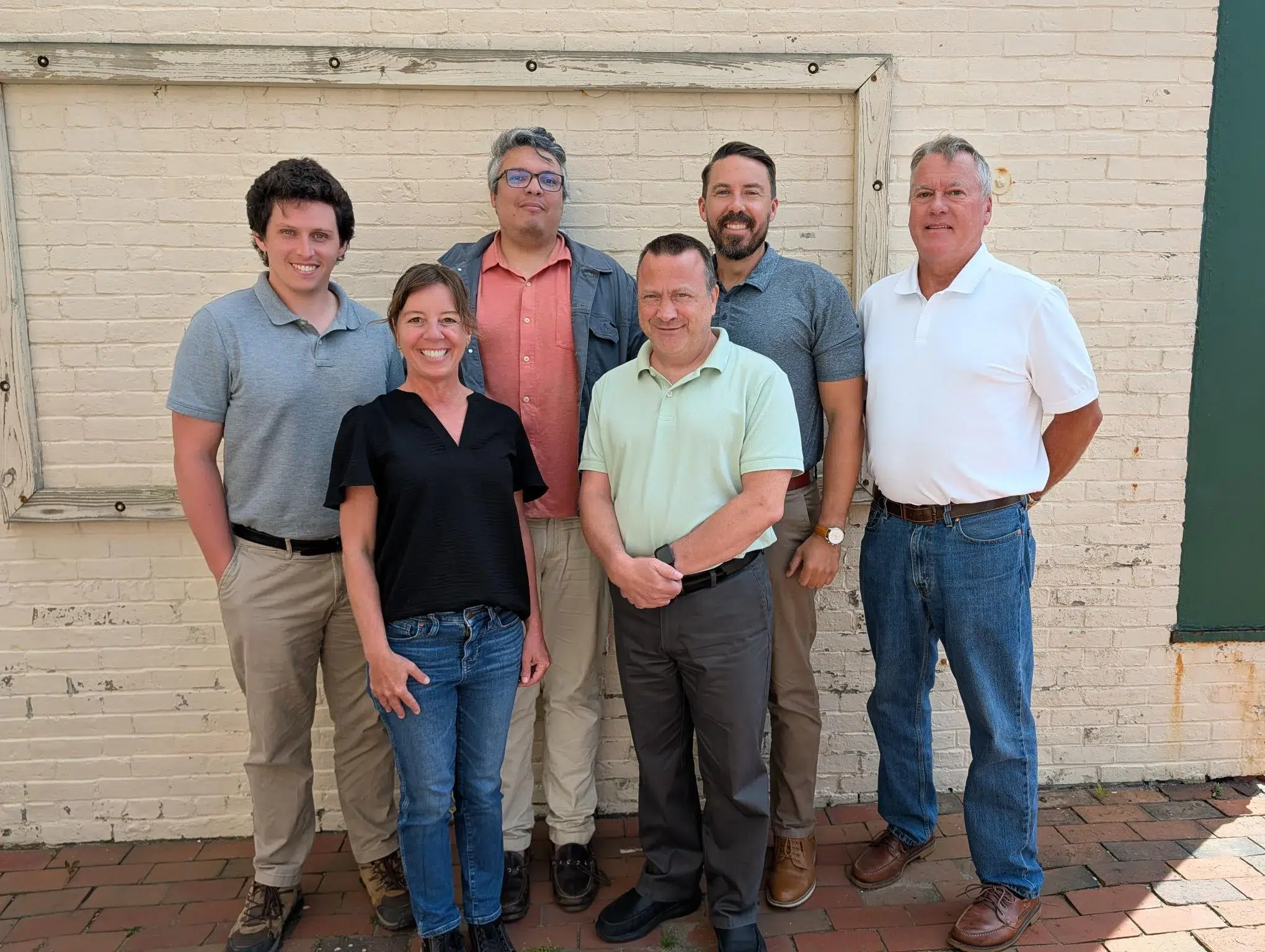 Seven adults posing together outdoors in front of a light brick wall with a wooden frame. They wear casual shirts and jeans and smile at the camera.