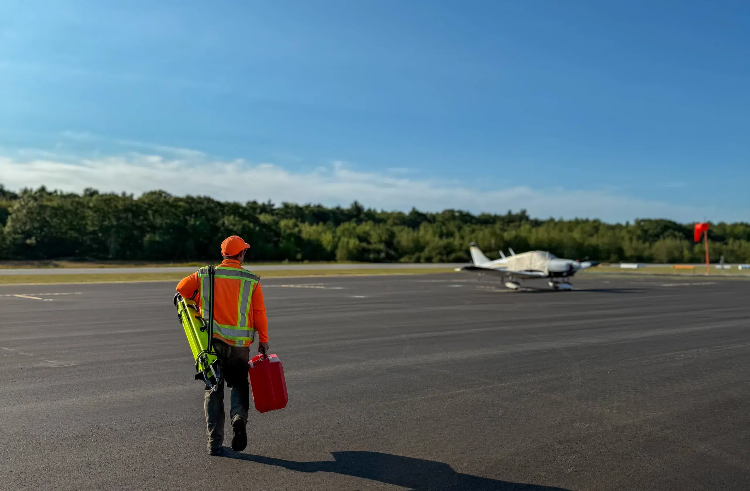 A person in a safety vest and orange hat carries a fuel can toward a small airplane on an airport tarmac, possibly as part of projects managed by civil engineering companies.
