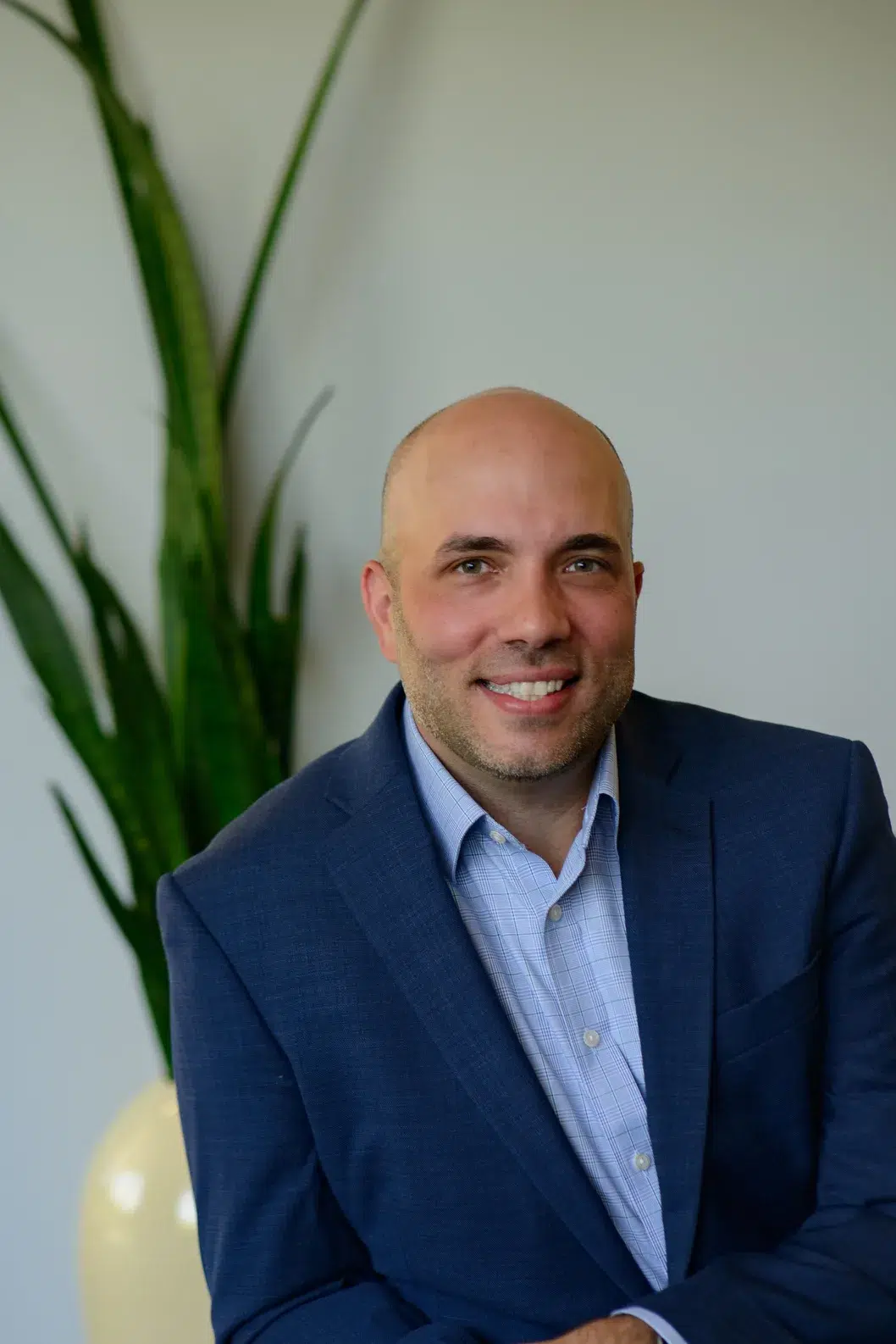 A smiling man in a blue blazer and light shirt sits in front of a tall green plant, with a neutral background.