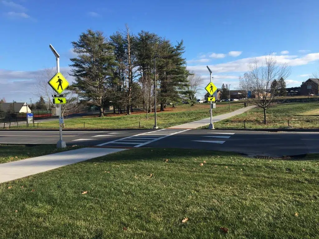 COLBY COLLEGE ATHLETIC COMPLEX - 1 - Sebago Technics A crosswalk with pedestrian crossing signs, designed with expert civil engineering services, connects a sidewalk to a path through a grassy park with trees under a blue sky.