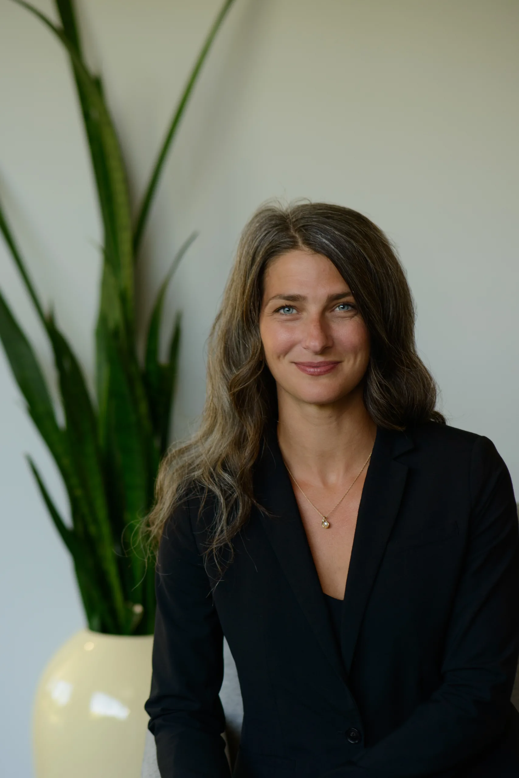 A woman with long wavy hair wearing a black blazer sits in front of a large green plant in a cream-colored pot, smiling slightly.