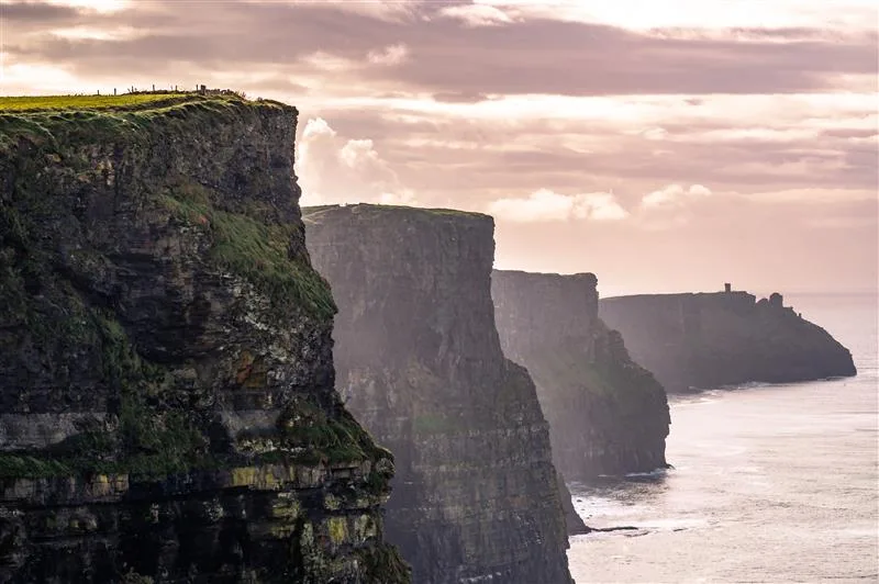 Cliffs of Moher - Sebago Technics Dramatic sea cliffs rise above the ocean under a cloudy sky, with green grass atop and people visible along the edge—an inspiring view for any Maine engineering firm.