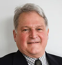 Smiling older man with gray hair, wearing a dark suit jacket, checkered shirt, and dark tie, representing a Maine engineering firm against a plain light background.