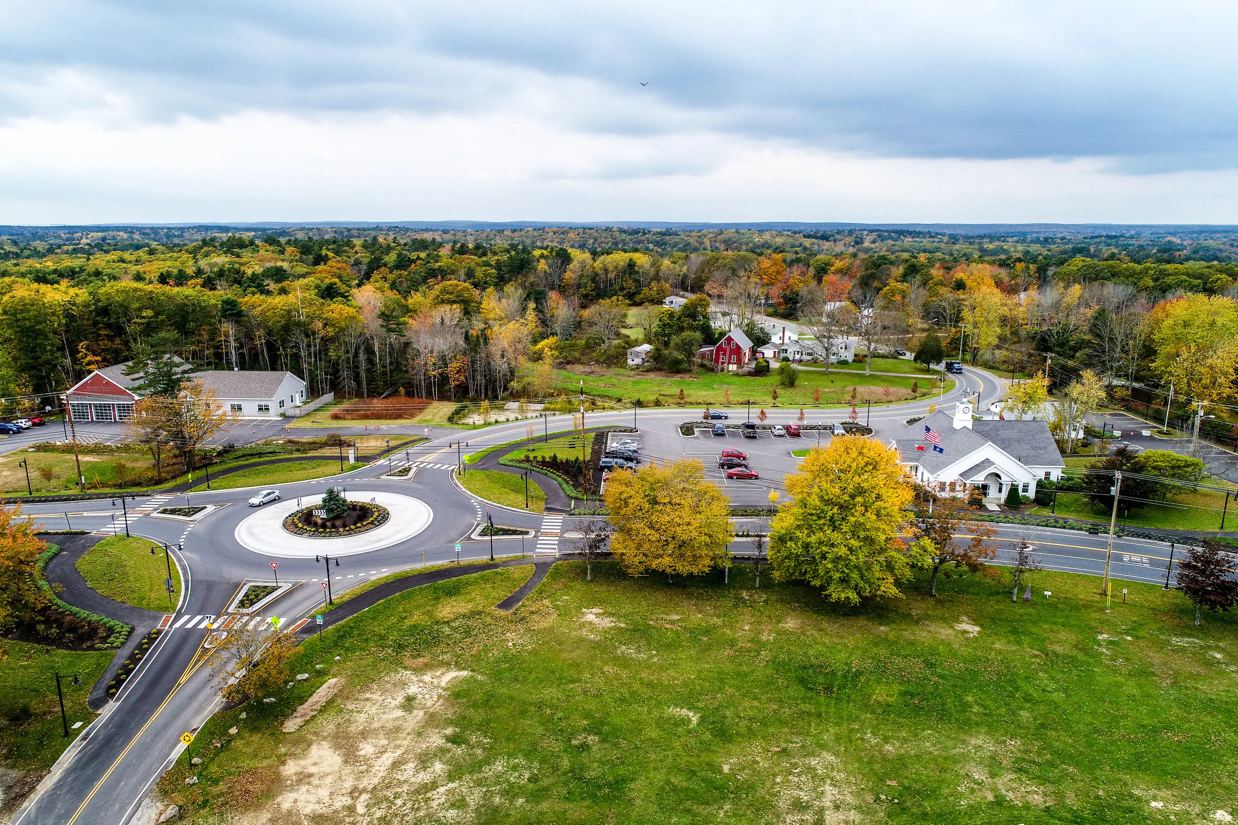 DJI_0073 (1) - Sebago Technics Aerial view of a roundabout, a hallmark of civil engineering, surrounded by autumn trees, buildings, parking lot, and roads in a suburban area under a cloudy sky.