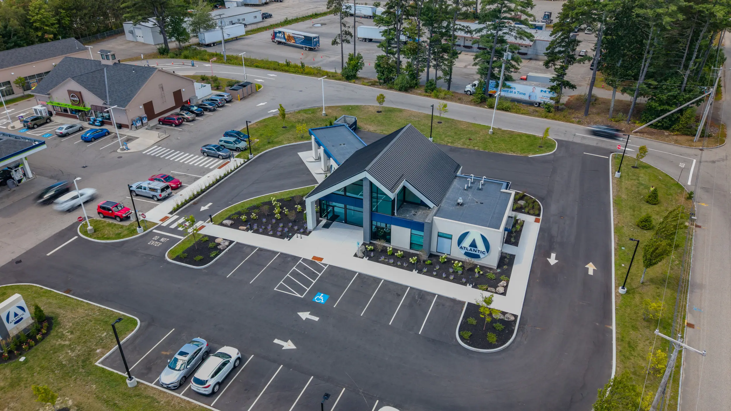 Aerial view of a modern, standalone building offering civil engineering services, with a blue and white logo, surrounded by trees, parking lot, cars, and trucks nearby.