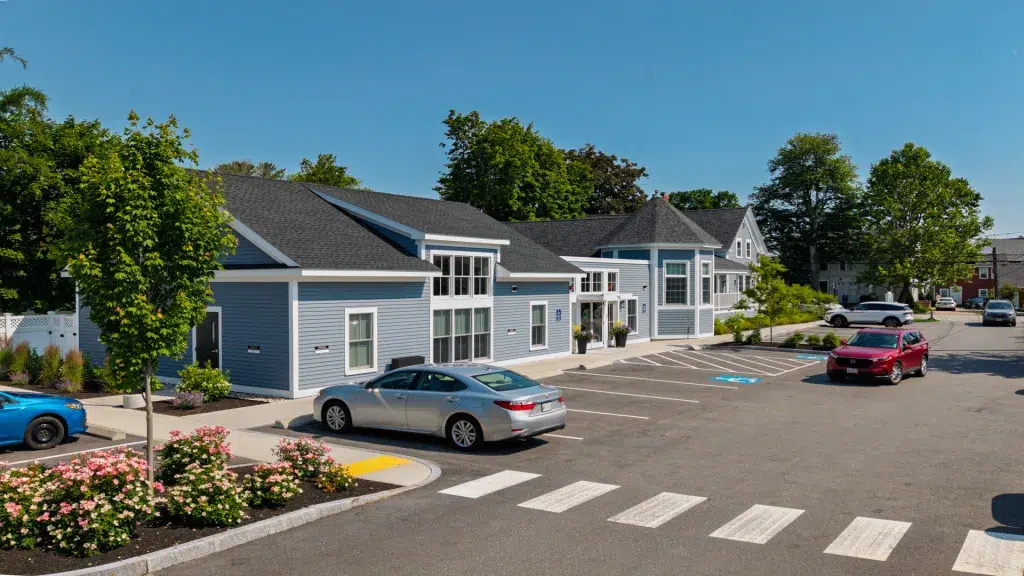 Blue residential complex with white trim, landscaped front, and a parking lot in the foreground under a clear sky.