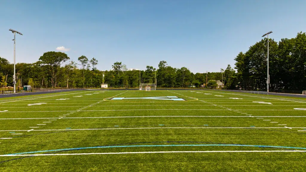 default - Sebago Technics Wide view of an empty football field with goalposts in the distance, white yard lines, and surrounding track under a blue sky.