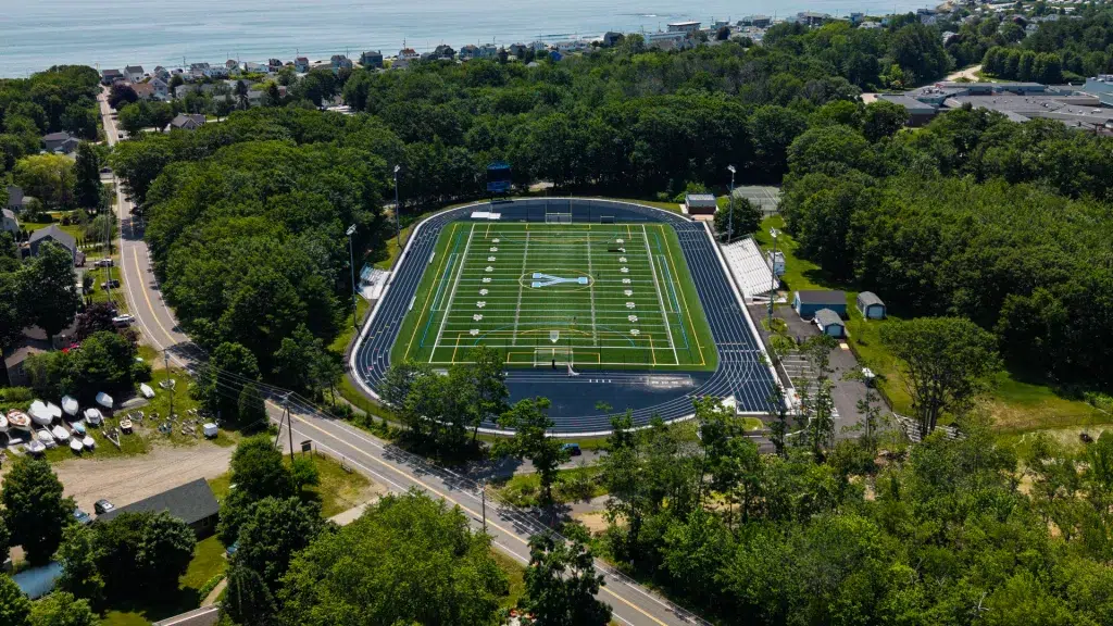 Aerial view of a football field inside a blue-track oval, surrounded by trees and a coastal town.