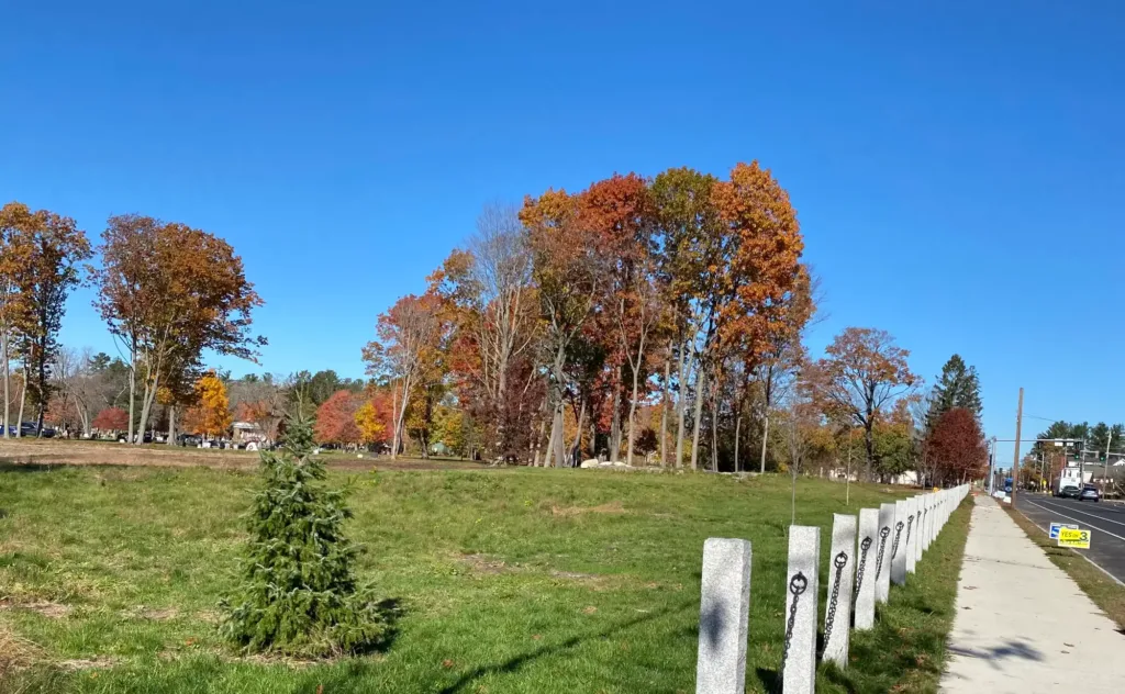 A sidewalk runs beside a grassy field and a row of posts, with colorful autumn trees under a clear blue sky.