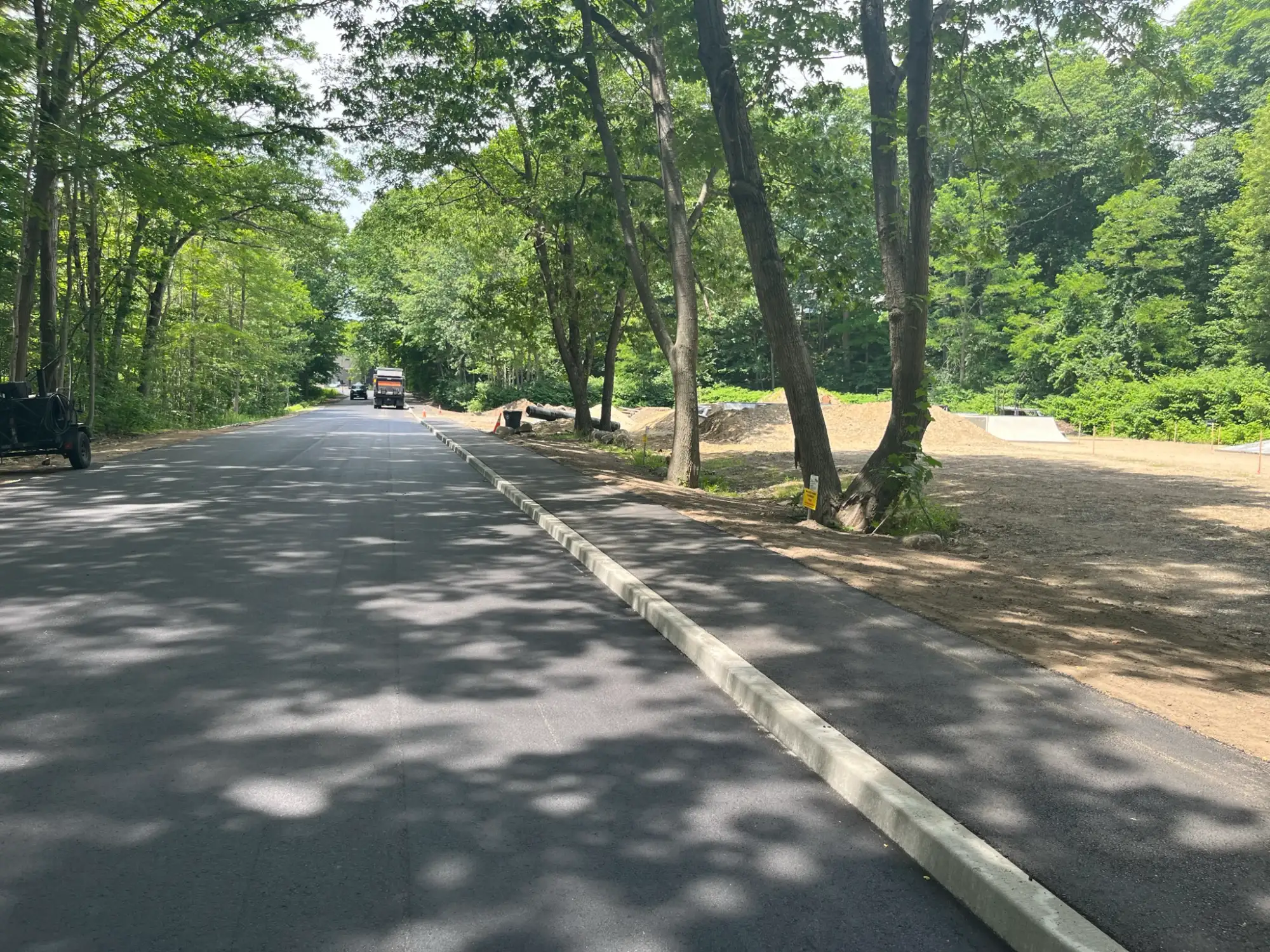 A newly paved road with fresh curbs runs through a wooded area; construction equipment and cleared land are visible on the right side.