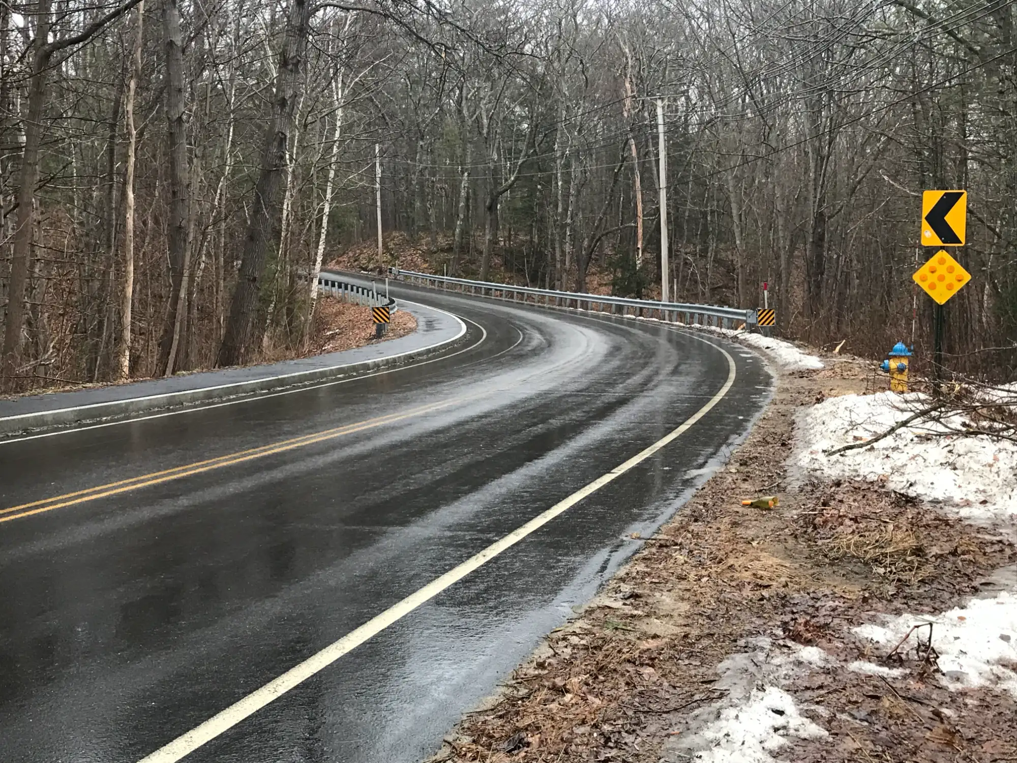 FACTORY PASTURE LANE SIDEWALK 2 (1) - Sebago Technics A wet, winding road curves through a forest with bare trees, snow patches on the ground, and yellow road signs warning of the sharp turn ahead.