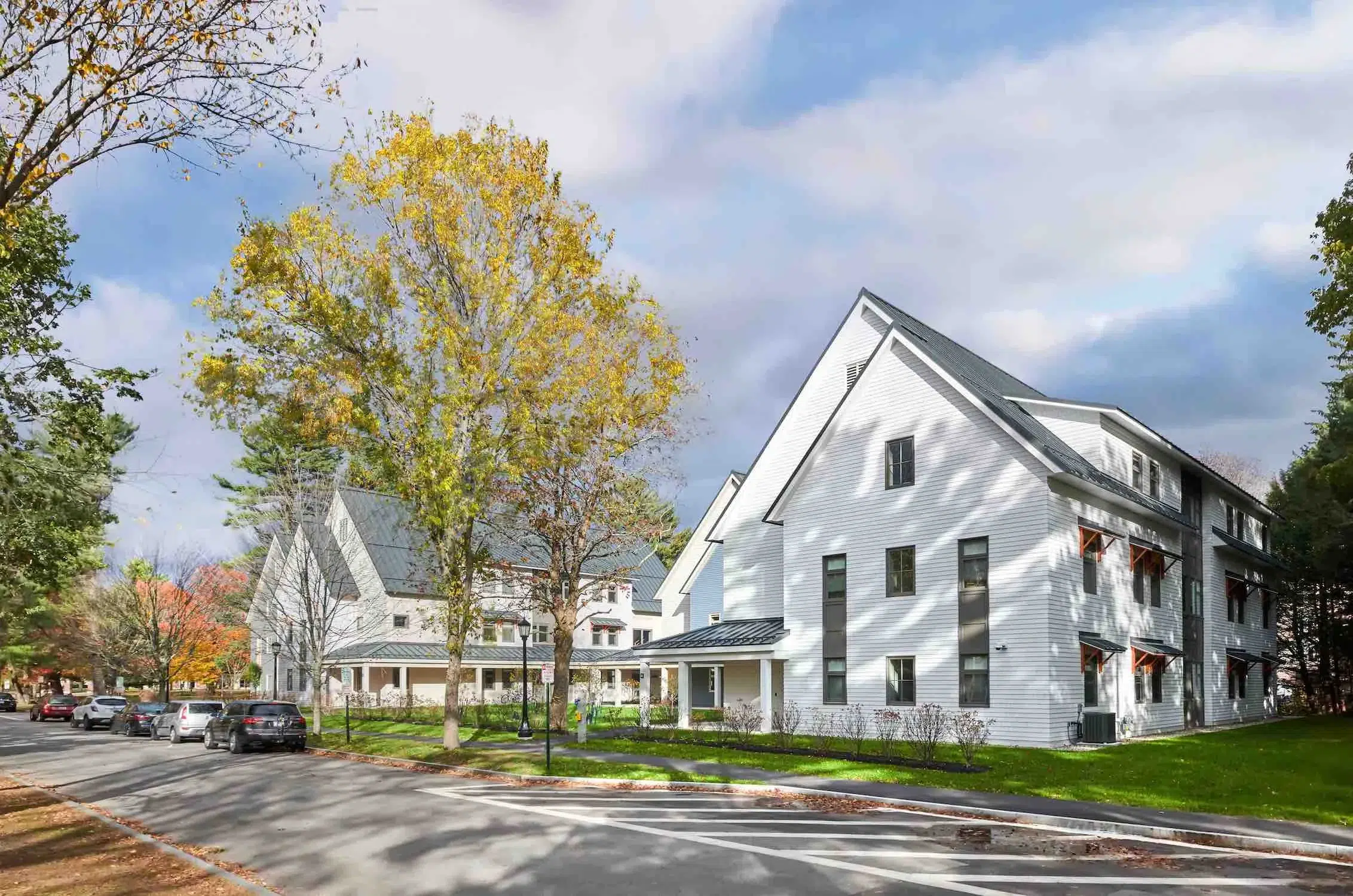 A white, modern three-story building with large windows, surrounded by trees with autumn leaves, is located along a quiet street with parked cars.