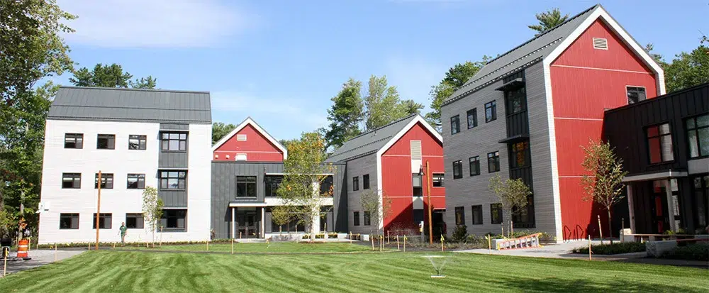 HARPSWELL AND PARK ROW APARTMENTS - Sebago Technics Three modern multi-story buildings, with red and white exteriors and large windows, surround a green lawn under a sunny sky with scattered clouds.