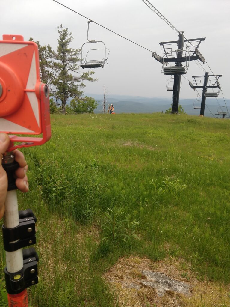 A person from a Maine engineering firm holds surveying equipment on a grassy slope near ski lift chairs, with mountains in the background under a cloudy sky.