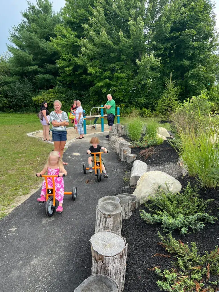 Two young children ride orange tricycles on a paved path outdoors, while adults and kids—perhaps from a Maine engineering firm outing—gather in the background by trees and greenery.