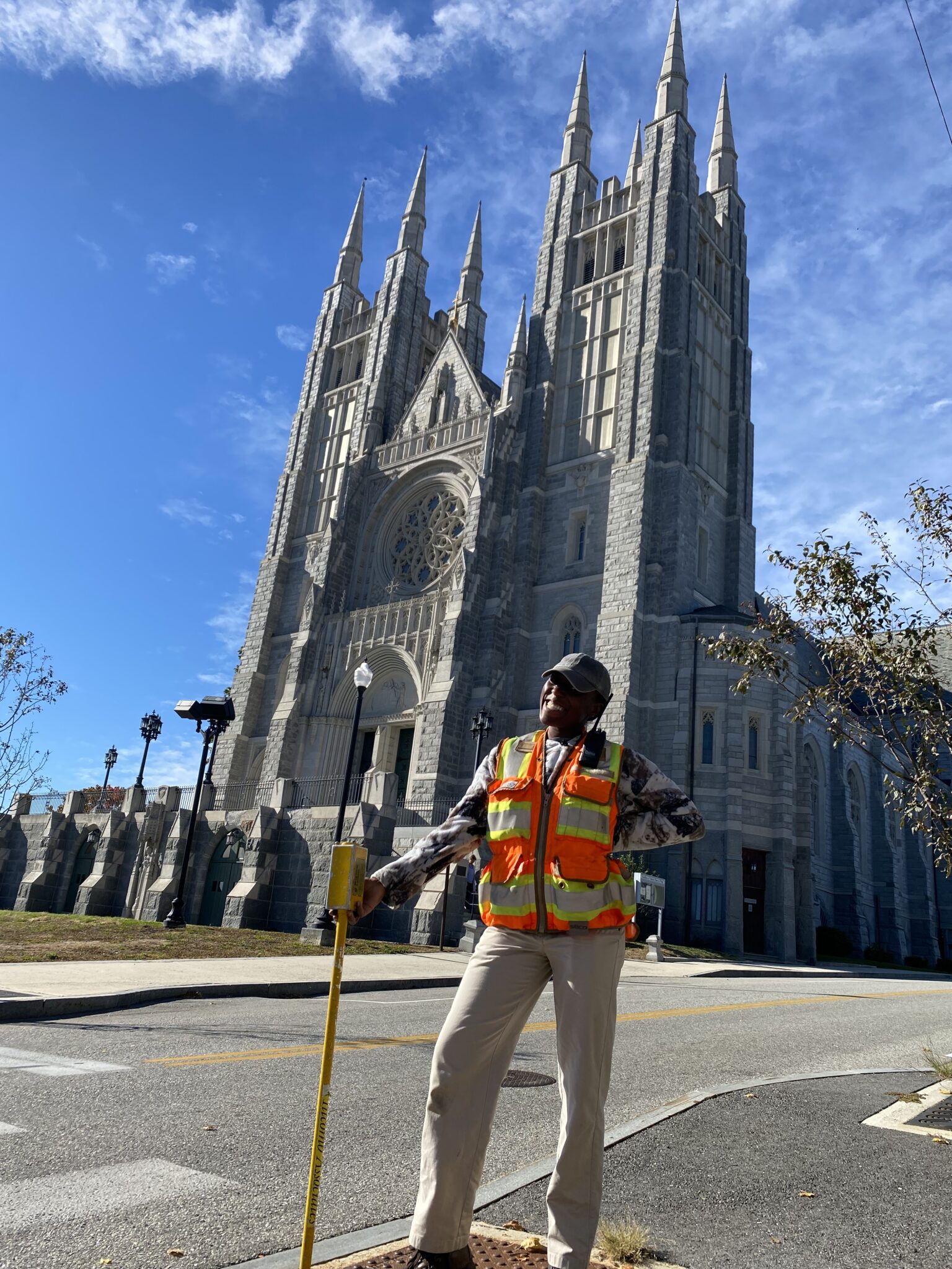 A person in a safety vest and hard hat, from a Maine engineering firm, stands by a crosswalk button with an ornate church behind them under a clear blue sky.