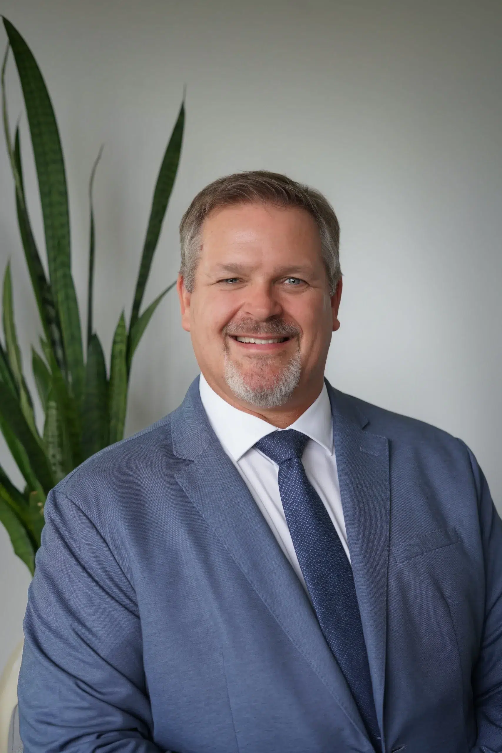Smiling man in a blue suit and tie, with a tall green plant in the background indoors.