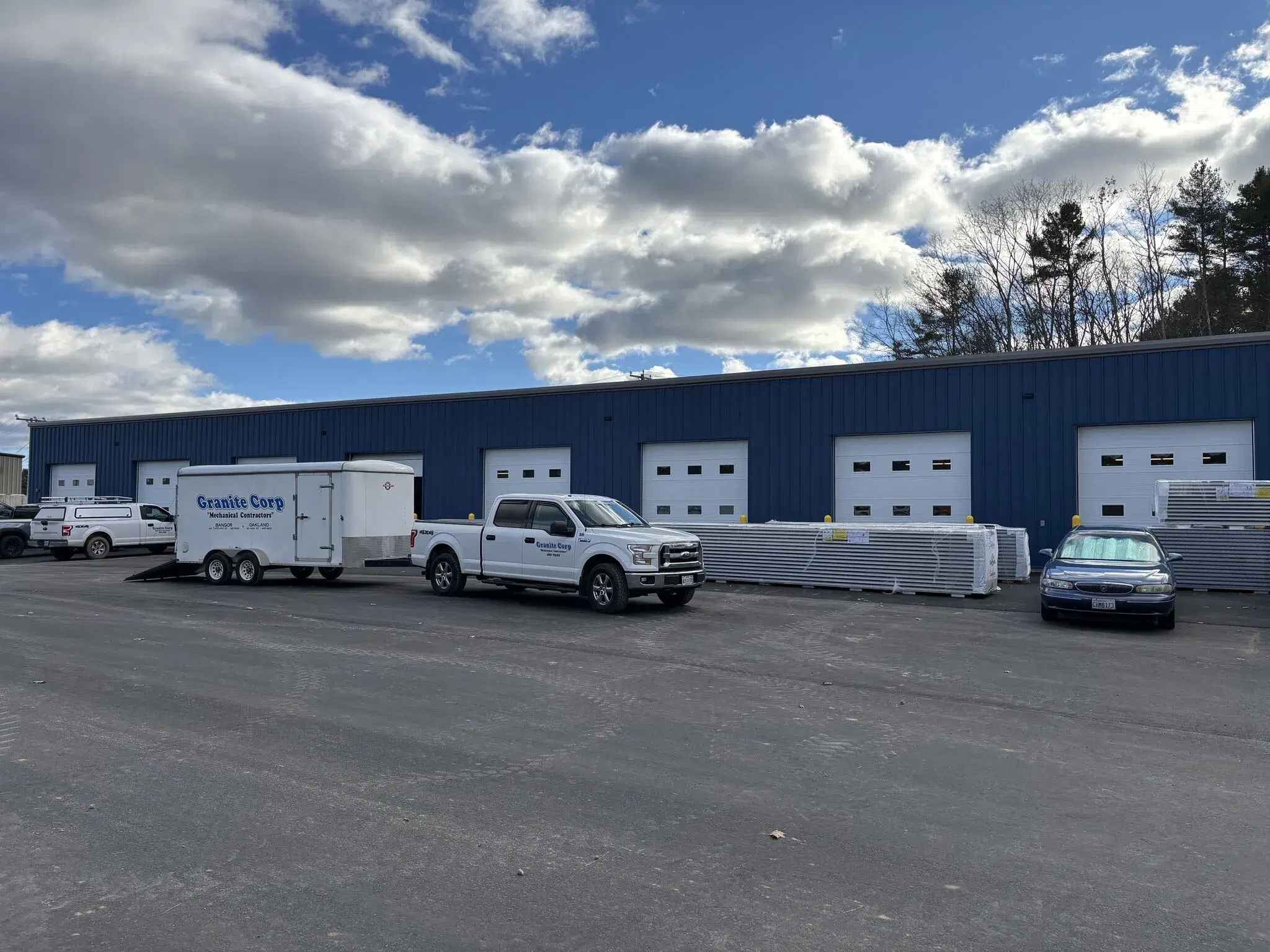 KENNEBUNK PUBLIC WORKS FACILITY 2 - Sebago Technics A blue industrial warehouse with multiple garage doors, several parked vehicles, a white trailer, and stacks of building materials outside on a sunny day.
