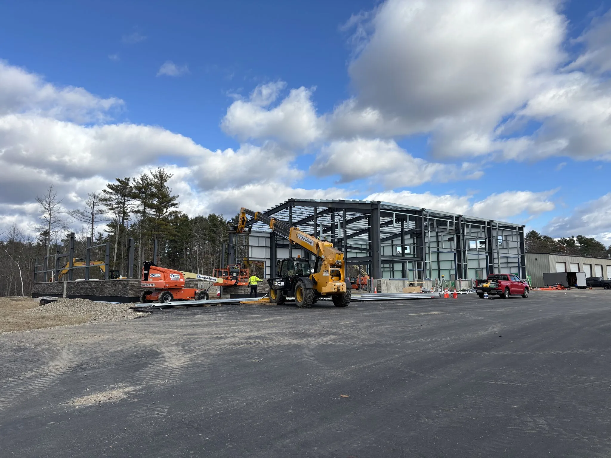 Construction site with workers and vehicles building a large steel-framed structure under a partly cloudy sky, surrounded by trees and construction equipment.