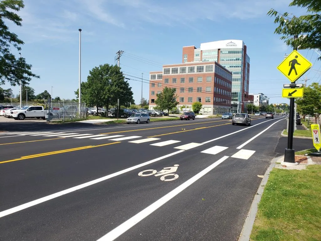 A freshly paved road with bike and pedestrian lanes, crosswalks, cars parked and driving, and a large office building in the background.