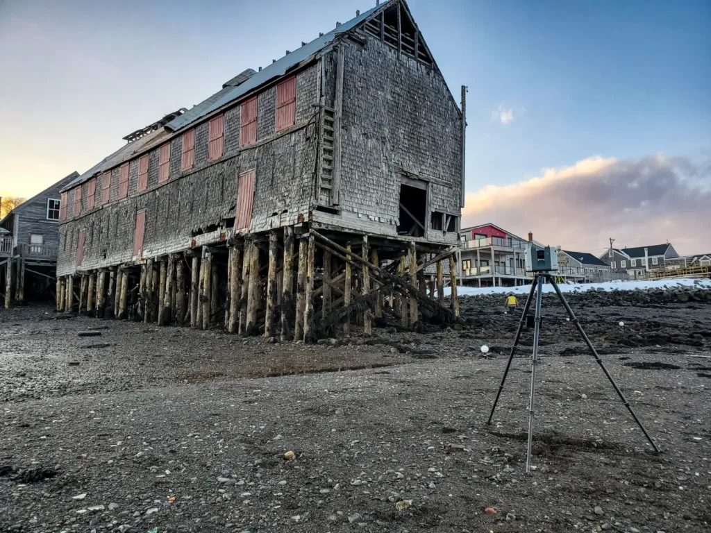 A weathered wooden building on stilts stands on a rocky shore at low tide, with houses and a camera tripod visible in the background at sunset.