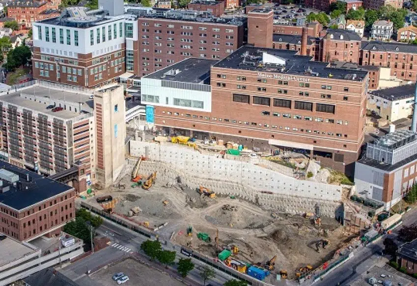 MMC Master Planning - Sebago Technics Aerial view of a large construction site by a multi-story hospital in an urban area, featuring dirt, machinery, and roads—planned by a leading Maine engineering firm.