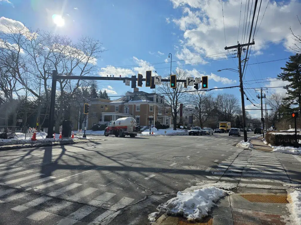 Intersection with traffic signals and crosswalks on a sunny winter day; snow along the curb and a tanker truck in the street.