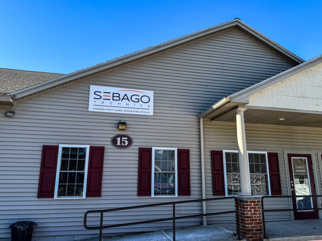 A tan building with red shutters displays a sign reading Sebago Technics, a Maine engineering firm, above the entrance. The number 15 is near the door, and a ramp leads up to the entrance.