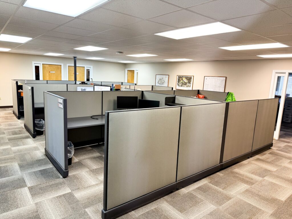 An office space at a Maine engineering firm with several empty cubicles, gray walls, computers, and chairs under bright fluorescent lights and a carpeted floor.