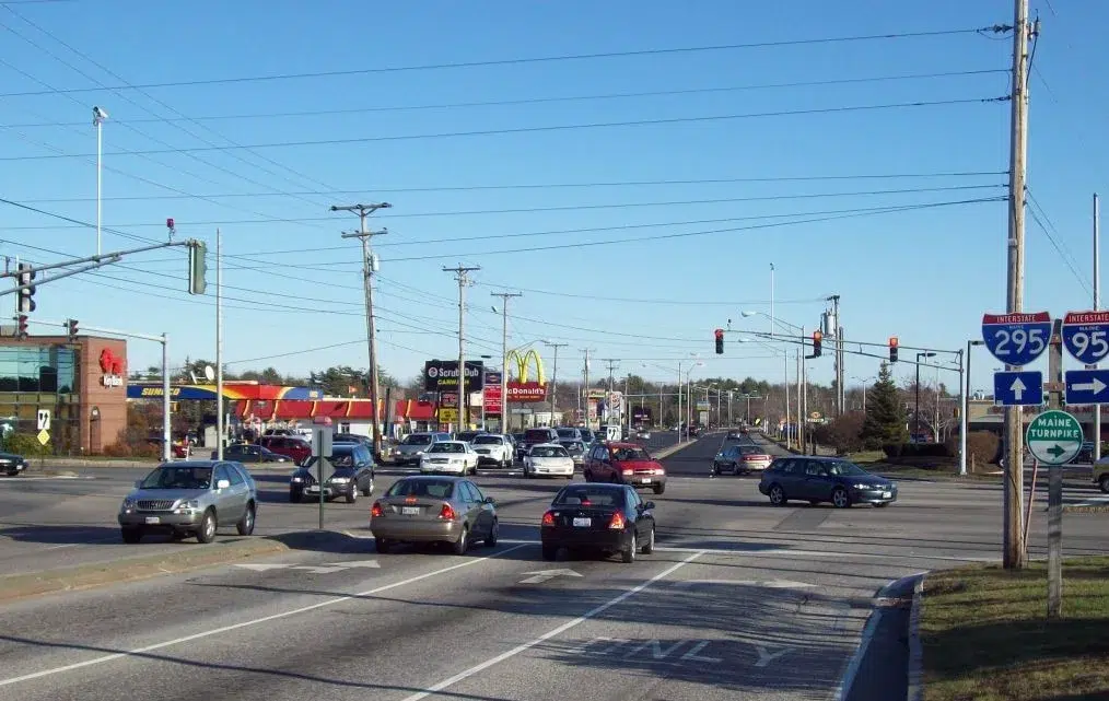 Cars wait at a busy intersection under a clear blue sky, with fast-food signs like McDonald’s visible and road signs for Interstate 295 and the Maine Turnpike on the right.