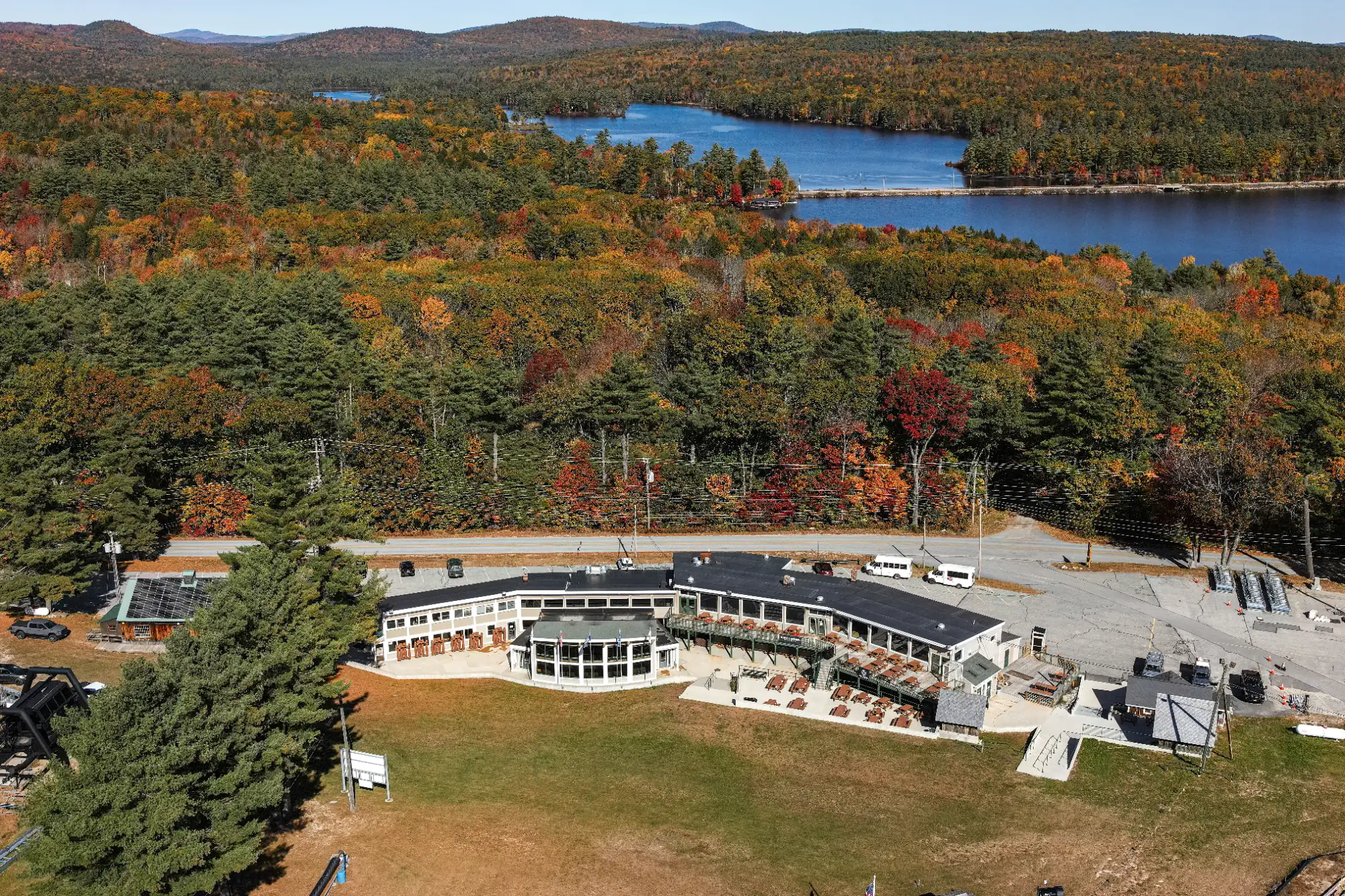 PLEASANT MOUNTAIN 2 - Sebago Technics Aerial view of a curved building surrounded by autumn trees near a lake, with hills in the background and a clear blue sky.