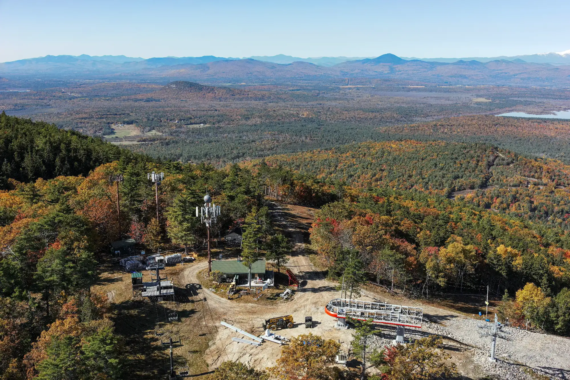 PLEASANT MOUNTAIN 3 - Sebago Technics Aerial view of a mountaintop construction site with equipment and ski lift, surrounded by autumn forest and distant mountains under a clear sky.