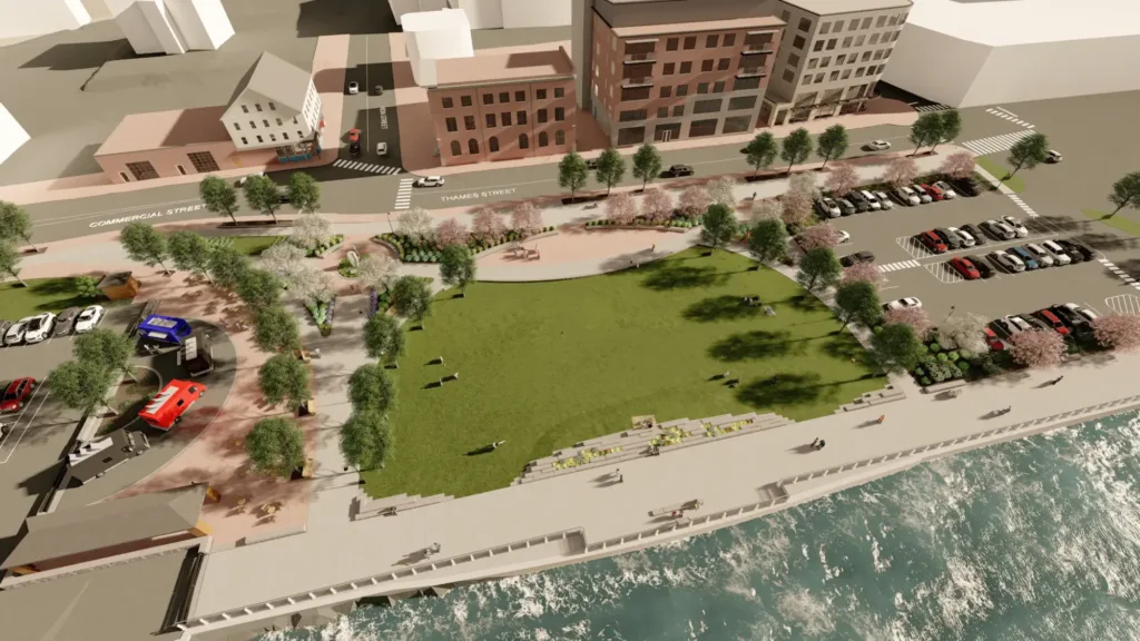 Aerial view of a waterfront park with a large grassy area, trees, walking paths, parked cars, nearby streets, and adjacent buildings.