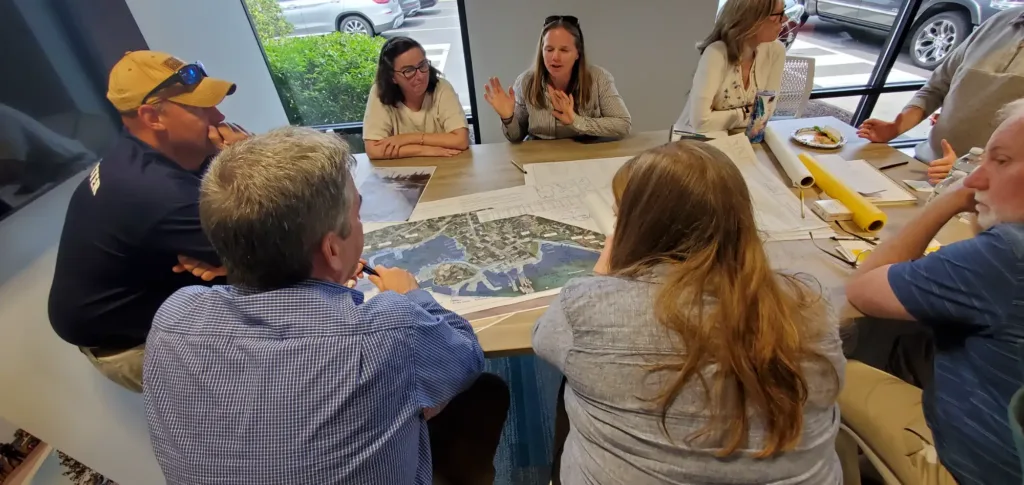 A group of people sit around a table discussing maps and documents in a bright meeting room, with windows showing parked cars outside.