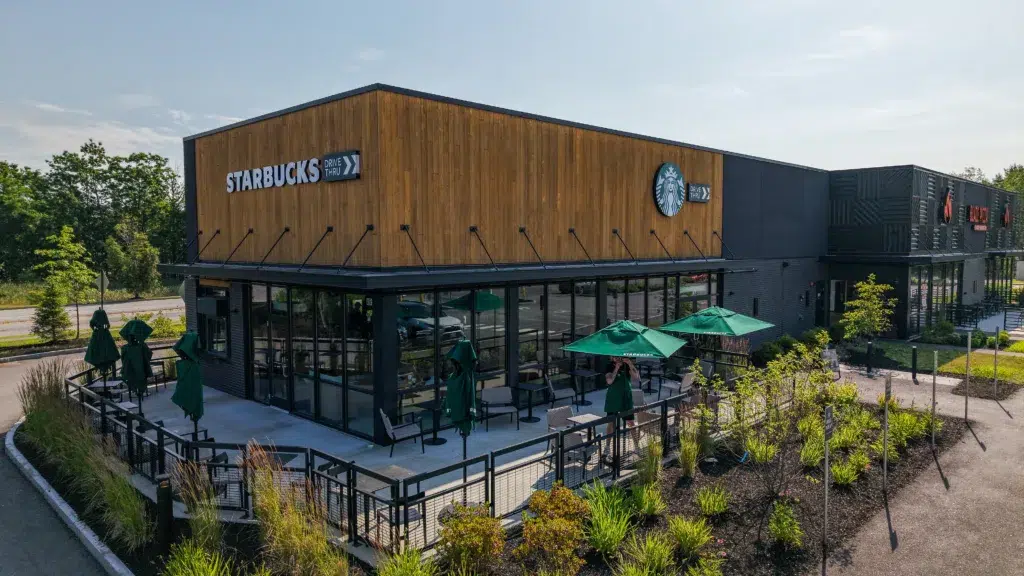 A modern Starbucks coffee shop with outdoor seating, green umbrellas, large windows, and wooden exterior accents, surrounded by landscaped greenery on a sunny day.