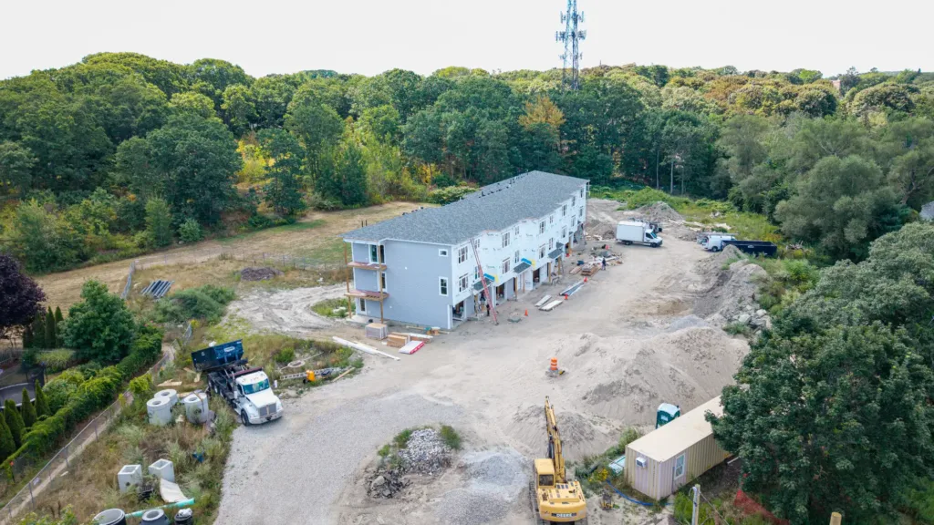 Aerial view of a construction site with a row of partially built townhouses, construction vehicles, equipment, and trees in the background.