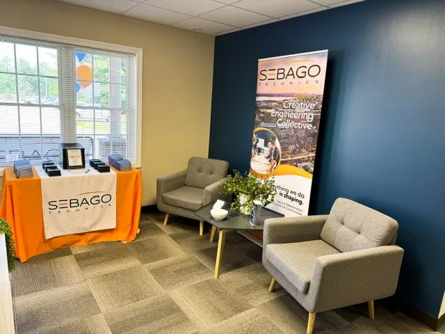 Sanford-Open-House-3 - Sebago Technics A modern office waiting area with two gray chairs, a plant, Sebago Technics banner, and brochures—reflecting the welcoming space of a Maine engineering firm.