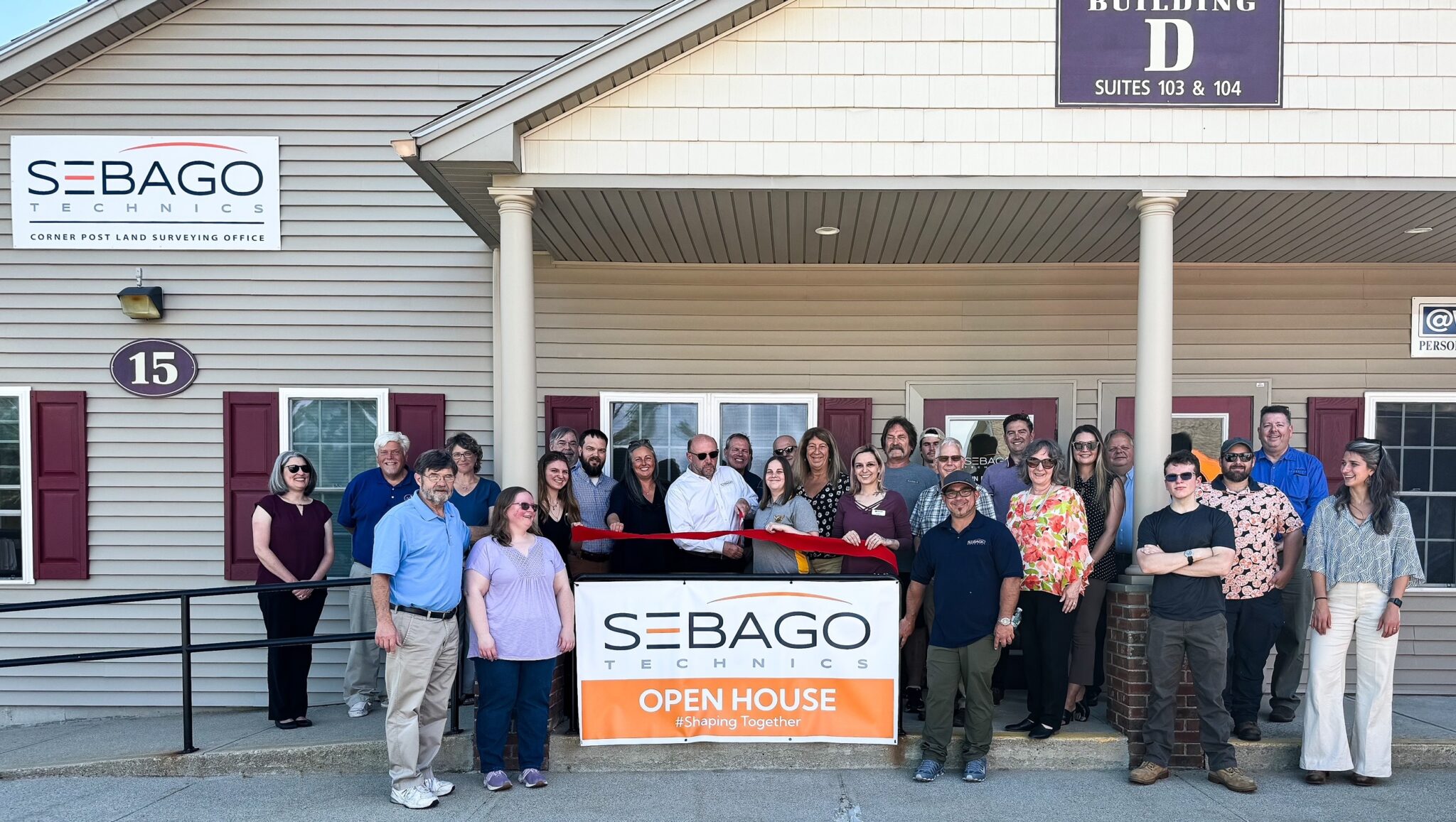 A group of people stand outside a building under a sign reading Sebago Technics, a Maine engineering firm, holding a ribbon and an Open House sign, smiling for a photo.