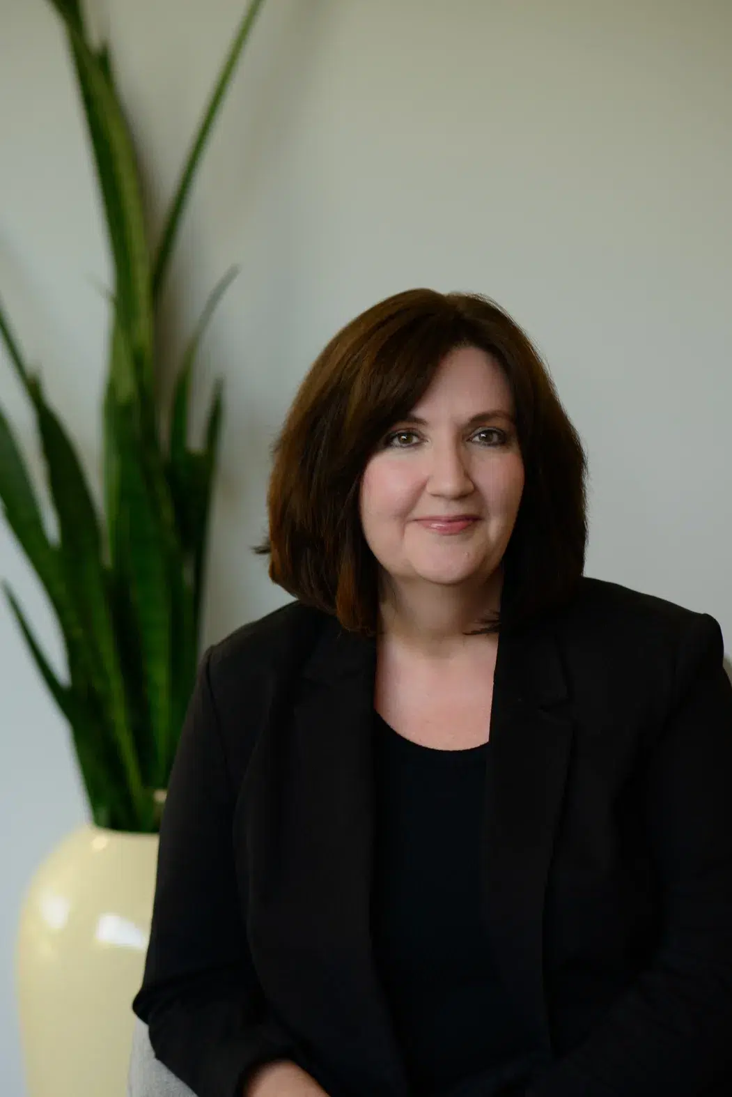 A woman with shoulder-length brown hair in a black blazer smiles while sitting indoors with a tall green plant in a white pot behind her.