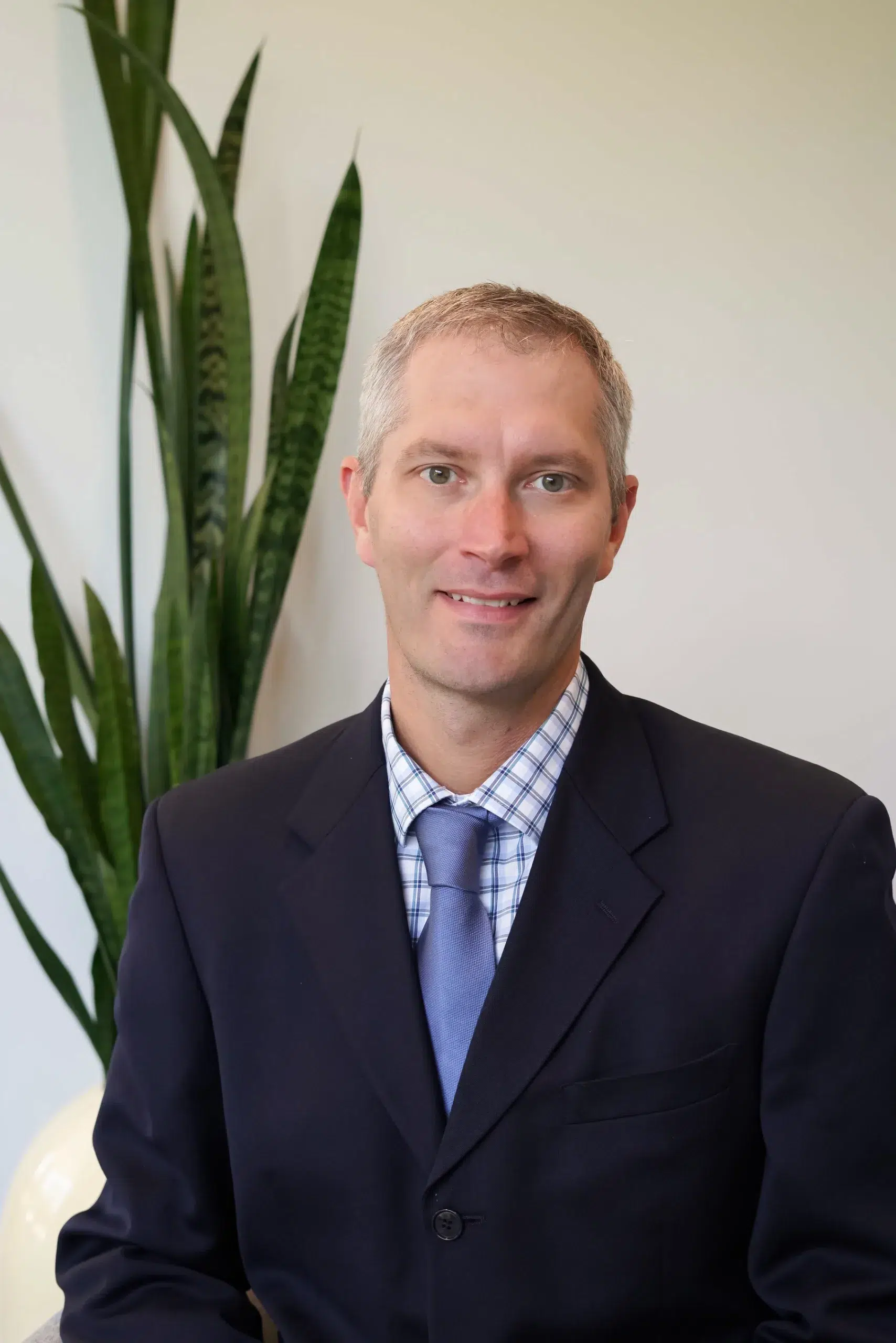 Head-and-shoulders portrait of a smiling man in a navy suit, light blue tie, and checkered shirt, with a tall plant in the background.