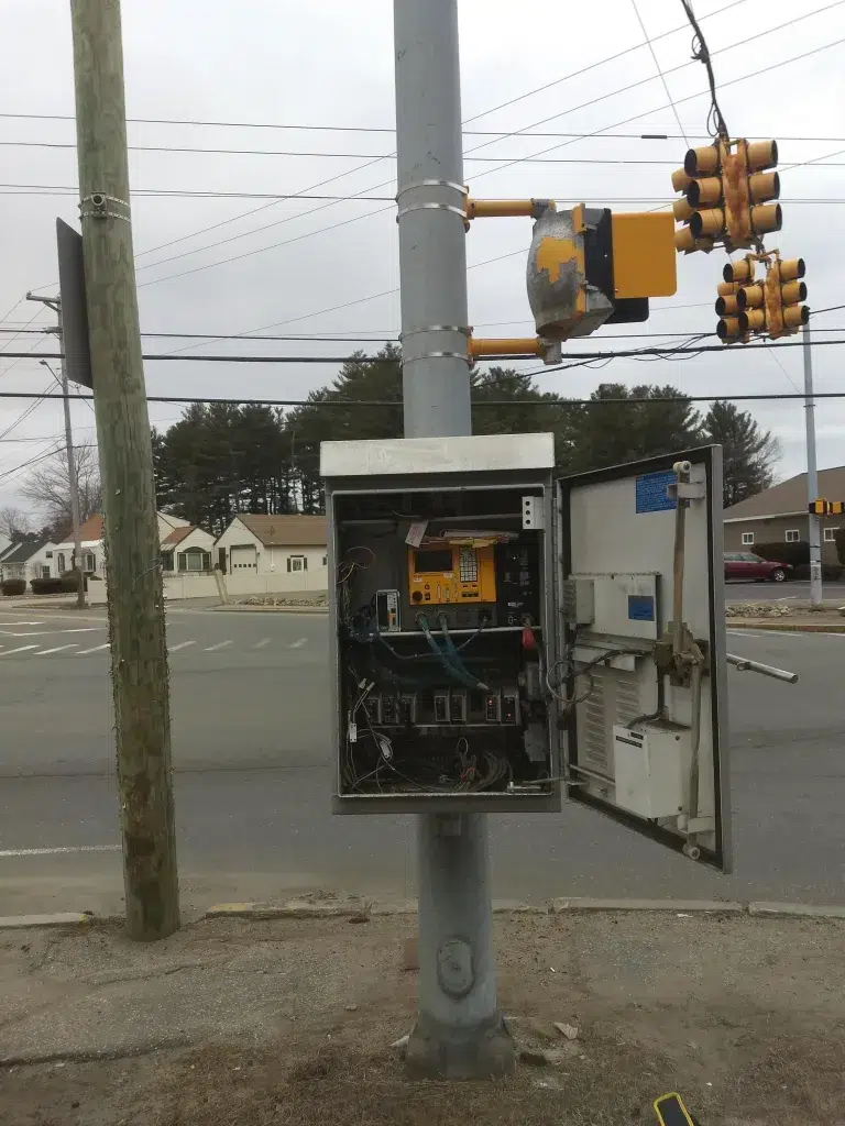 Somersworth High Street Corridor 1 - Sebago Technics Open electrical equipment cabinet mounted on a pole with exposed circuitry and wiring at a street intersection; yellow traffic lights visible in the background