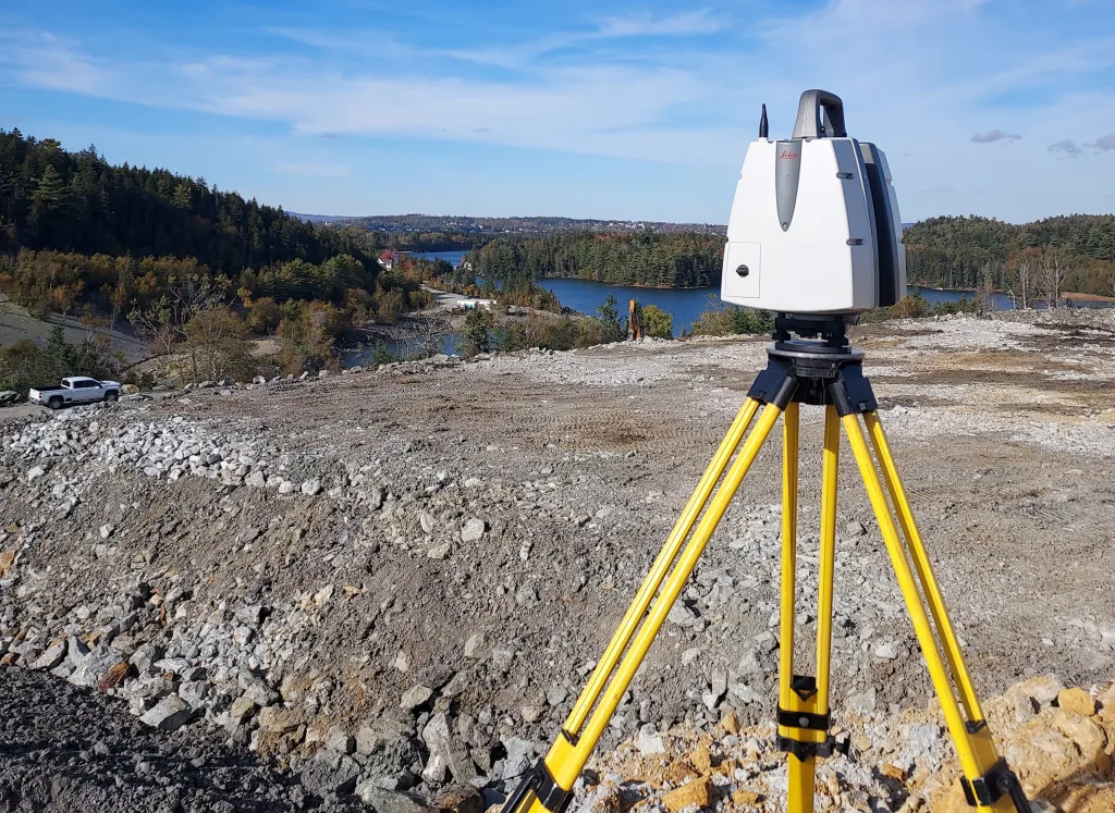 Surveying instrument (Leica total station) on a yellow tripod at a rocky construction site overlooking a lake and forested hills.