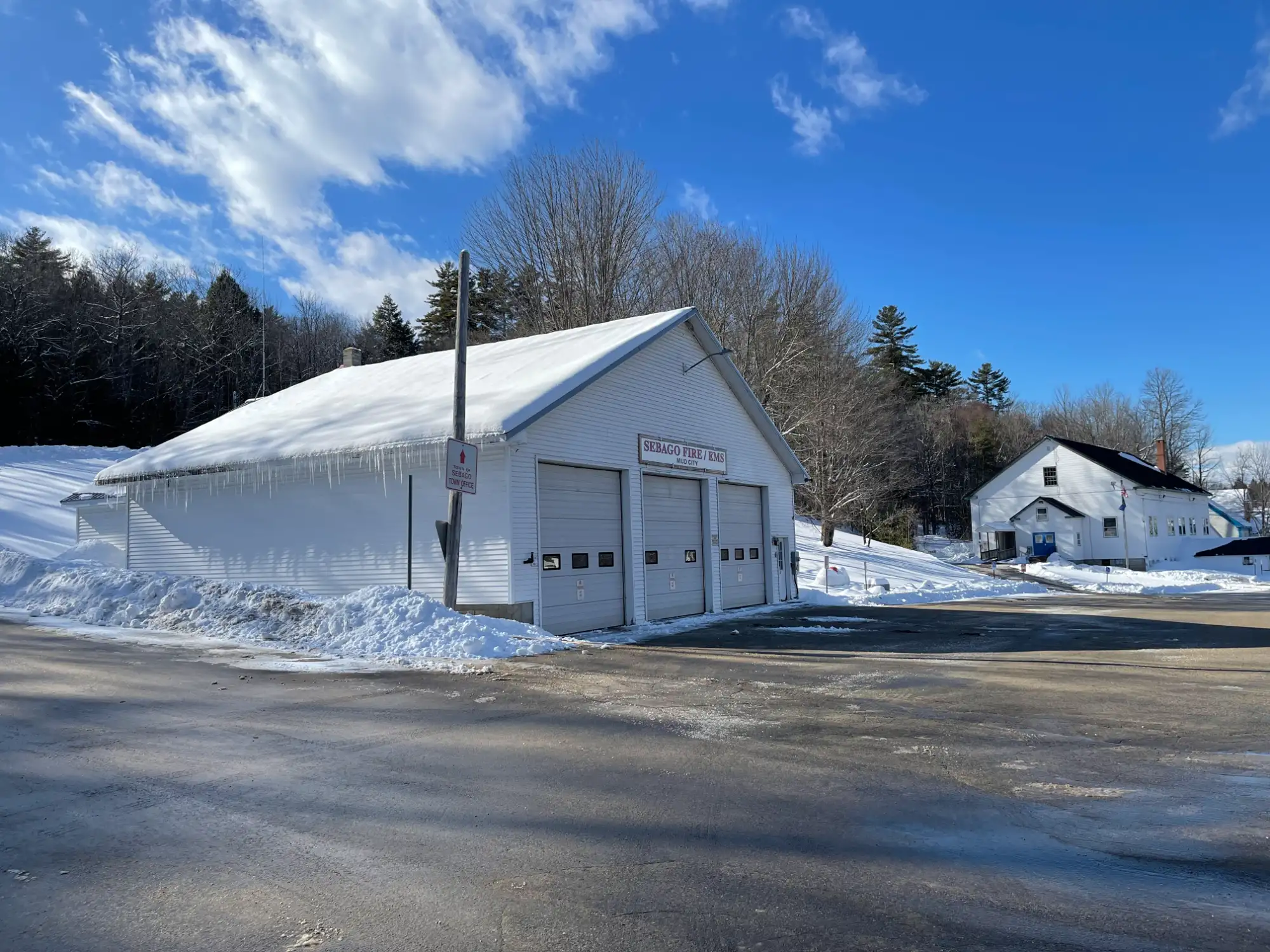 A white fire station with icicles hanging from the roof sits on a snowy lot under a blue sky, with a white house and trees in the background.