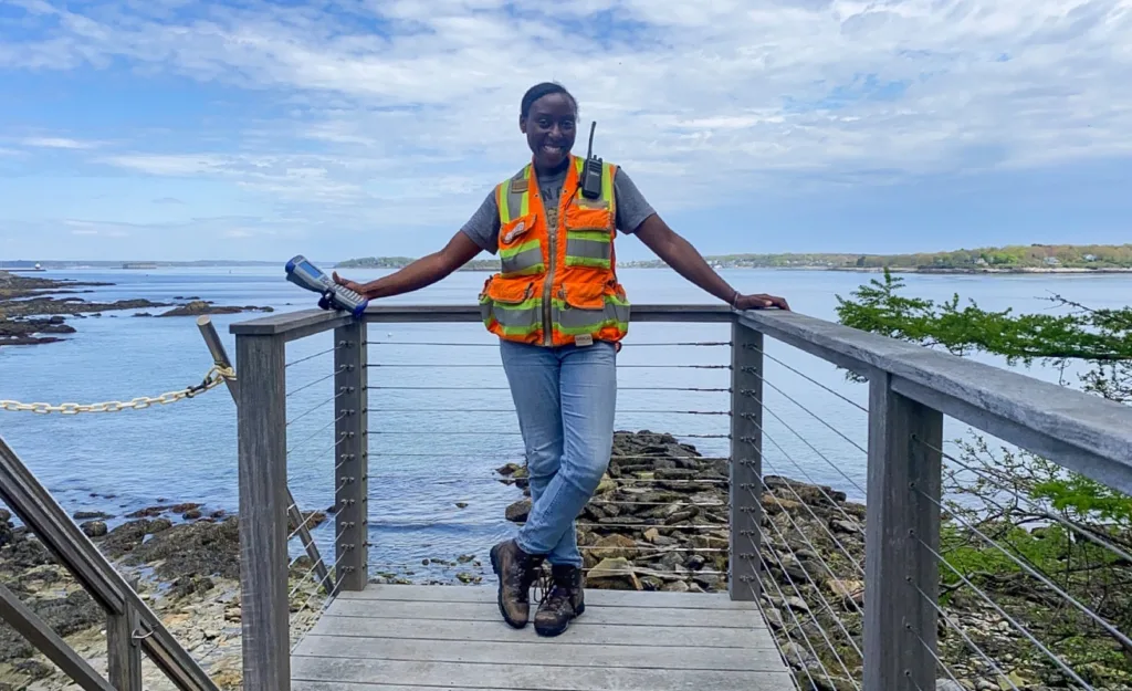 A person in a safety vest and boots stands smiling on a wooden platform by a rocky Maine shoreline, representing an engineering firm, with the ocean and blue sky in the background.