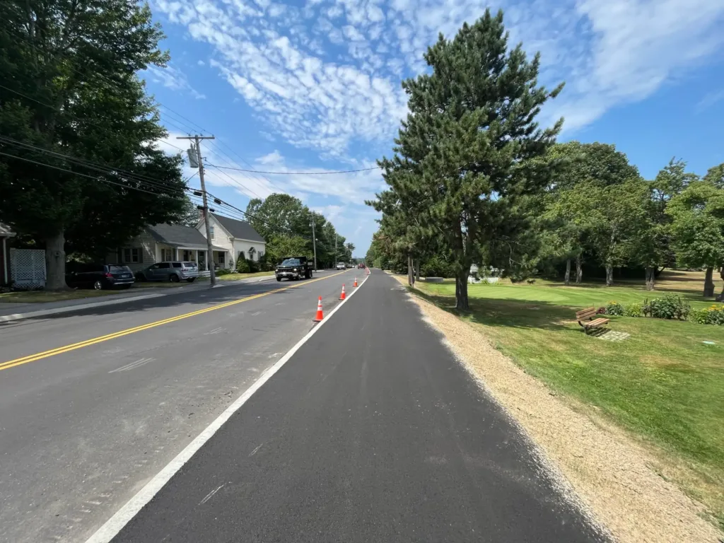 A newly paved road with orange traffic cones separating a bike lane from the main road, houses and trees line both sides under a blue sky with scattered clouds.